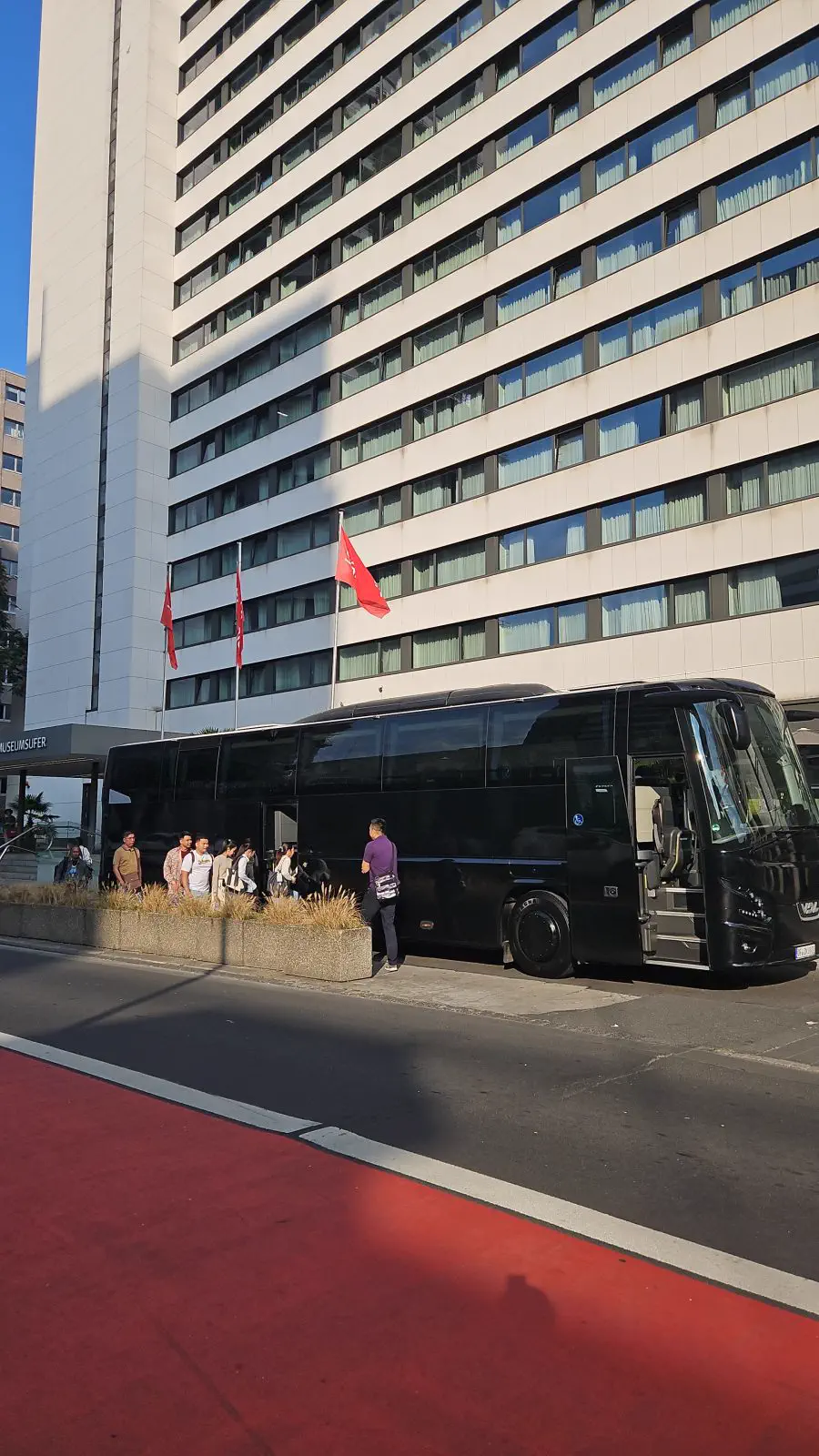 Schwarzer Reisebus hält vor einem hohen Hotelgebäude, Reisende stehen neben dem Bus