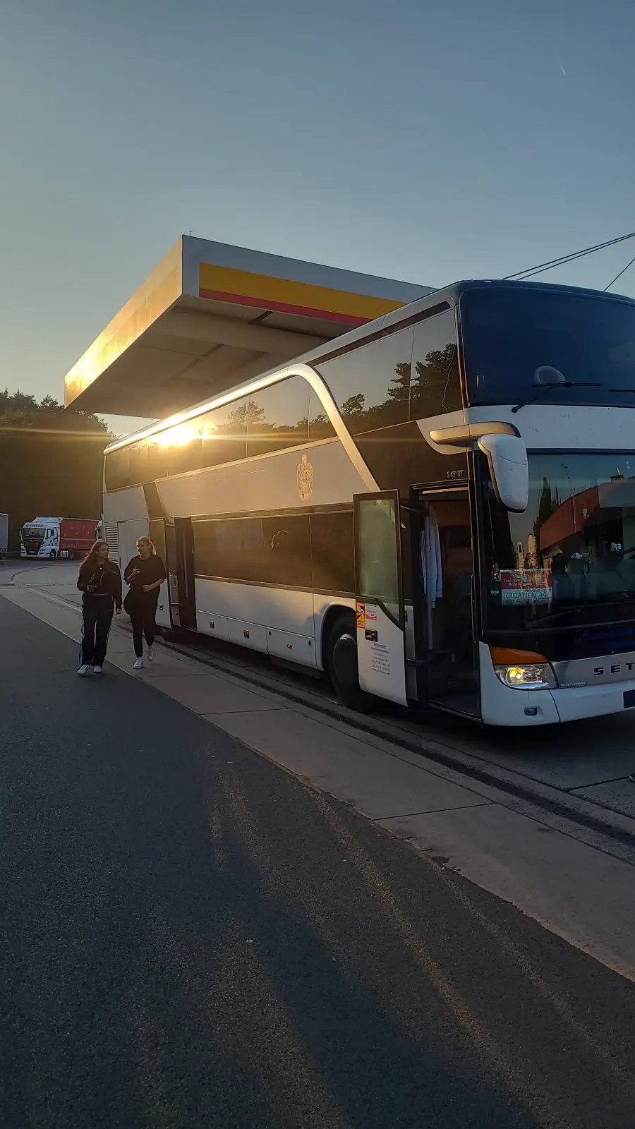 Setra-Doppeldeckerbus im Abendlicht an einer Tankstelle, zwei Reisende gehen vorbei